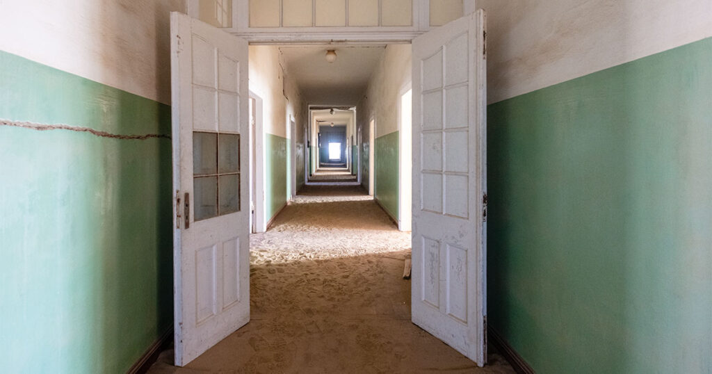 a hallway through a building with cracks in the plaster and light streaming through the doors on the  right side